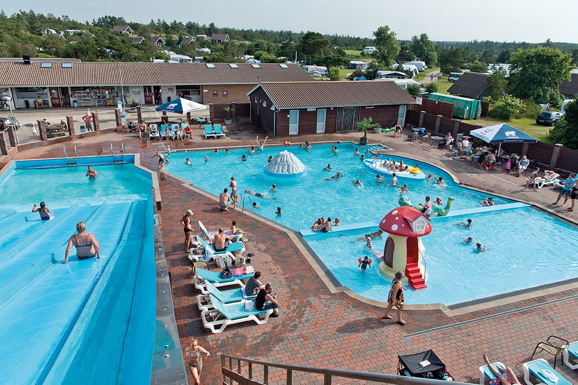 A family-friendly outdoor swimming area is depicted, featuring two pools interconnected by a slide. Various sun loungers and umbrellas are arranged around the pools, where individuals are enjoying the water. Colorful play structures provide entertainment for younger guests, and a backdrop of cabins and nature is visible.