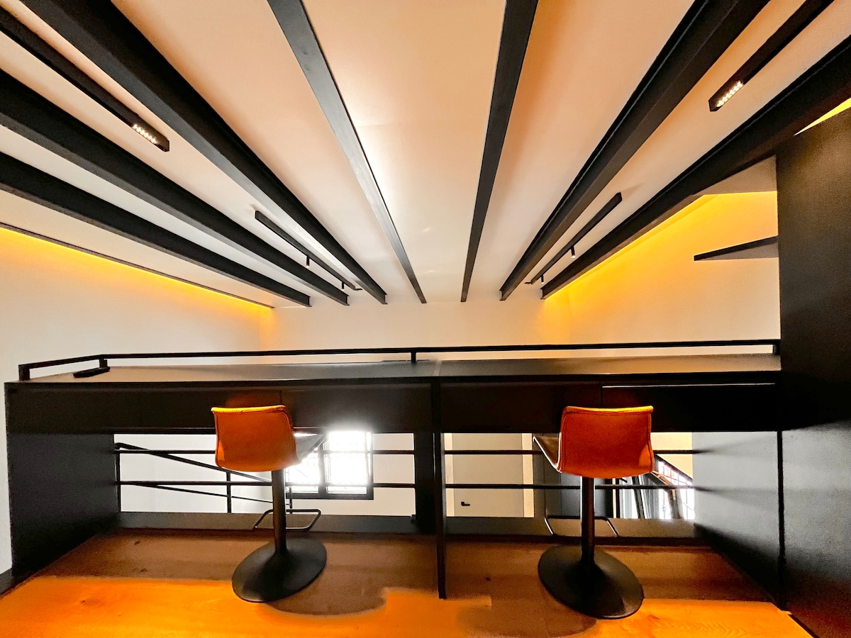 An upper-level work station is highlighted by sleek black railings and wooden flooring. Two modern stools are positioned at the desk, beneath a ceiling with linear lighting that provides a soft glow. The overall ambiance is clean and contemporary.