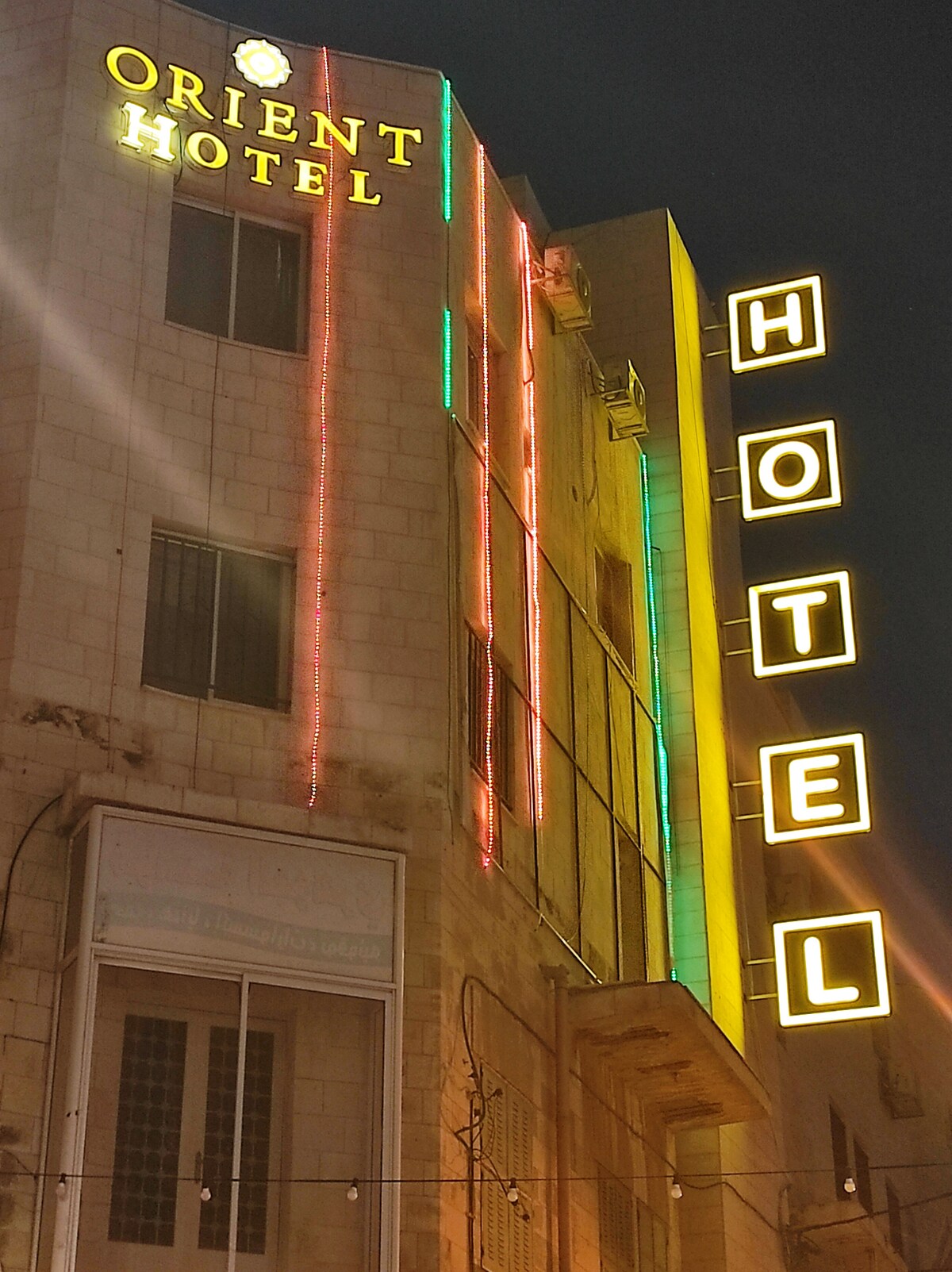 The hotel exterior is illuminated with vibrant neon signage, displaying 'Orient Hotel' and 'HOTEL.' The building features a blend of contemporary and traditional architecture, with stone walls and an array of colorful lights enhancing its nightlife ambiance.