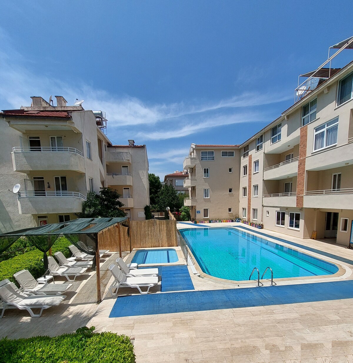 A well-maintained outdoor pool is highlighted in the center, surrounded by lounge chairs and greenery. Two multi-story buildings frame the pool area, with balconies visible from each unit, providing a view of the inviting pool and landscaped surroundings.