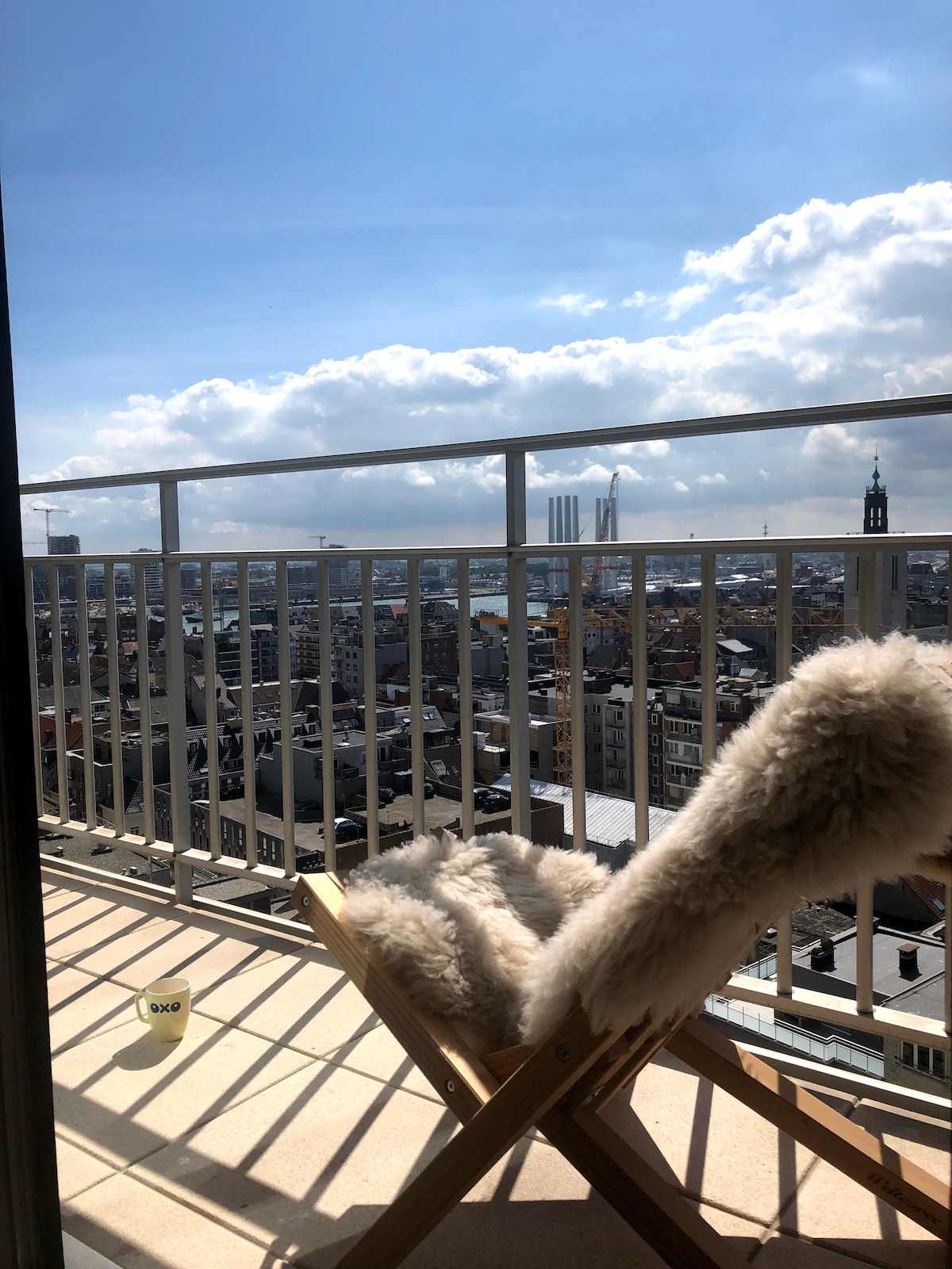 A view from a balcony is offered, showcasing an expansive cityscape under a partly cloudy sky. A wooden chair, draped with a soft, light-colored faux fur throw, is positioned on the terrace, inviting relaxation while gazing at the distant skyline.
