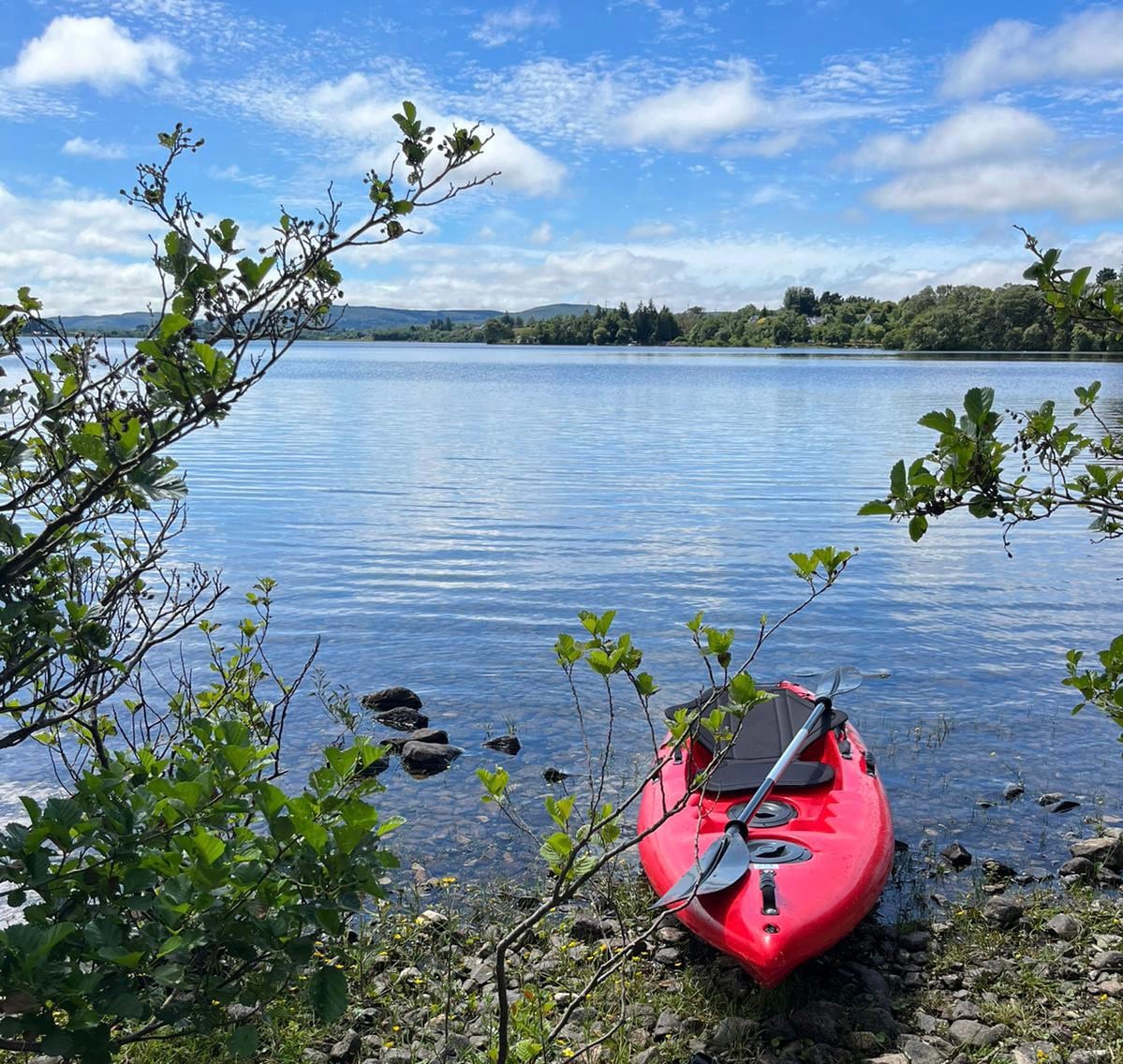 A red kayak is positioned on the rocky shore of a calm lake, surrounded by lush green foliage. Sunlight reflects off the still water, under a partly cloudy sky. The serene landscape offers a natural setting for watersports.