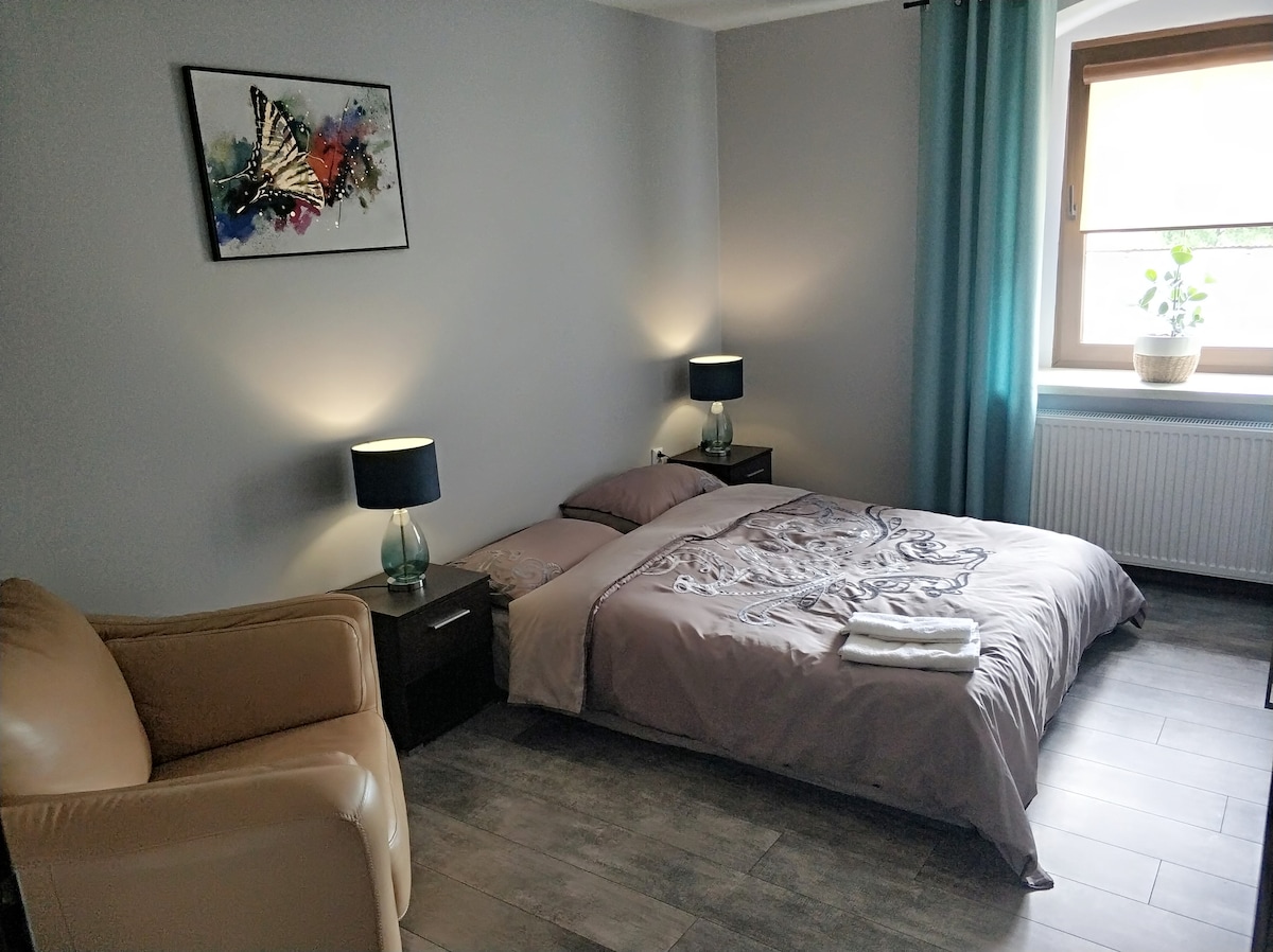 A sleeping area is observable, featuring a spacious bed with a decorative cover. Two bedside lamps provide soft lighting, while a comfortable armchair is positioned nearby. A window allows natural light to brighten the room, complemented by light blue curtains.