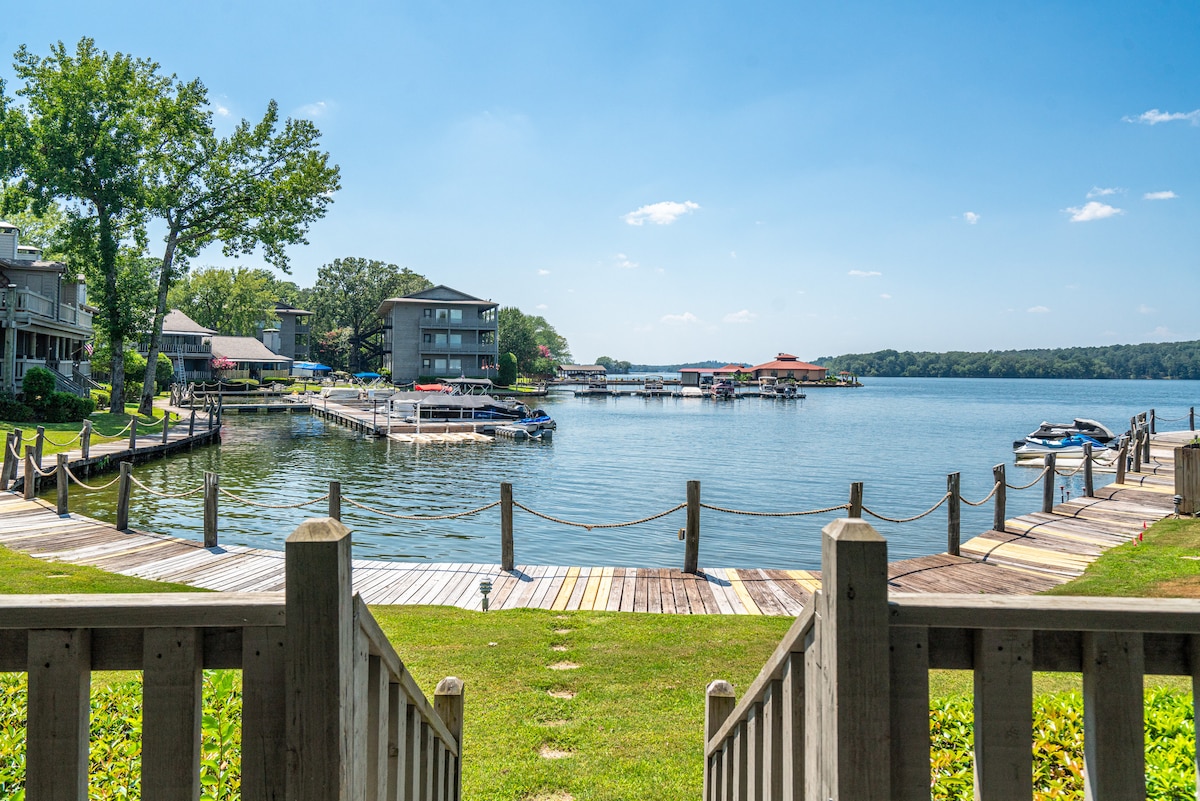 A serene lakefront view captures the calm waters of Lake Hamilton, framed by a wooden boardwalk and grassy areas. Multiple boats are docked alongside, with the distant shoreline dotted with vacation homes under a clear blue sky.