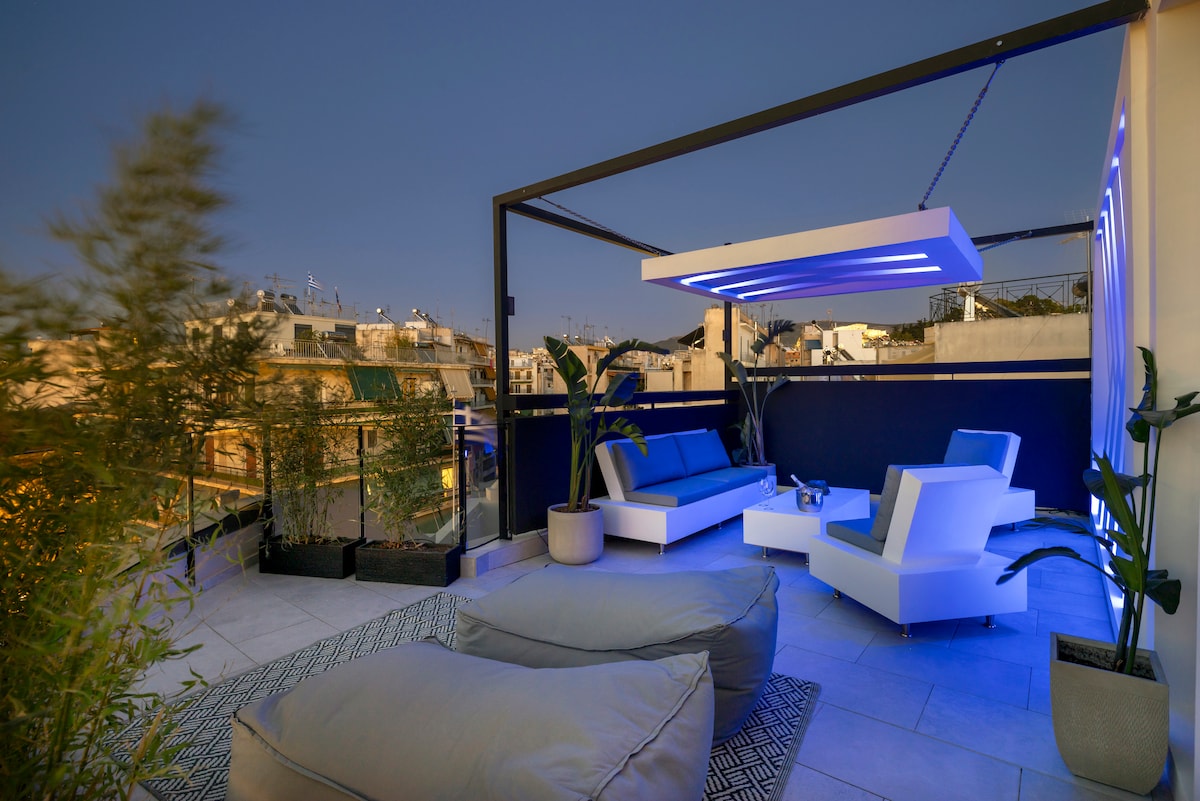 A private rooftop area is illuminated by soft blue lighting, showcasing a modern seating arrangement that includes a sleek couch and lounge chairs. Potted plants add a touch of greenery, and city views are visible beyond the railing, enhancing the relaxed outdoor ambiance.