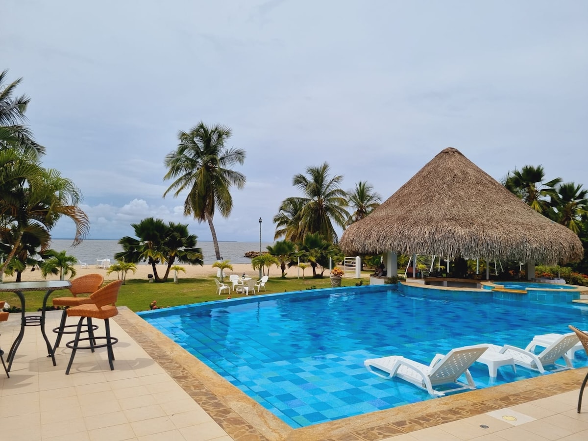A spacious outdoor area features a large pool with clear blue tiles that reflect the sky. Around the pool, lounge chairs and shaded seating under a straw-roofed gazebo are visible. Lush palm trees and beach views enhance the serene atmosphere.