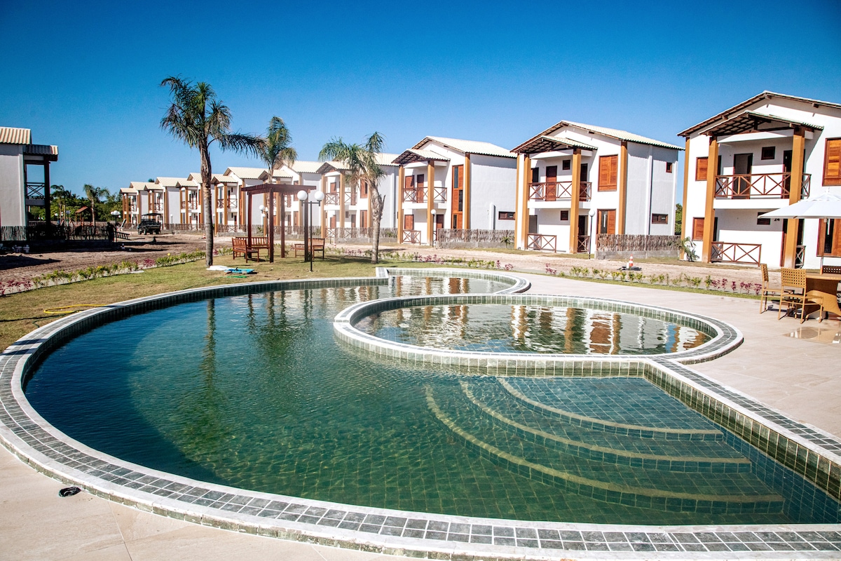 A large pool with a heart-shaped design reflects the clear blue sky. Surrounding the pool are several two-story buildings featuring wooden balconies and tasteful architecture. Lush greenery and palm trees enhance the outdoor space, creating a serene environment for relaxation.