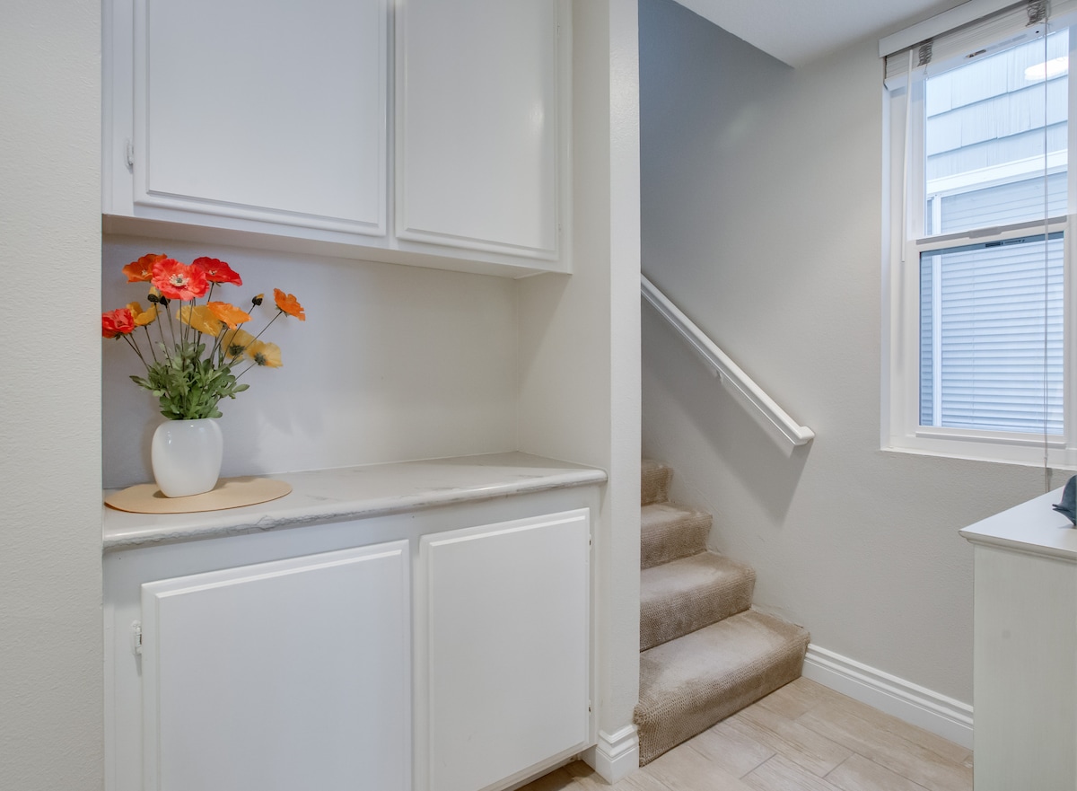 A well-lit entryway features a staircase leading to the upper level. A small countertop is adorned with a vase of colorful flowers, while white cabinetry provides storage options. Natural light filters in through the nearby window.