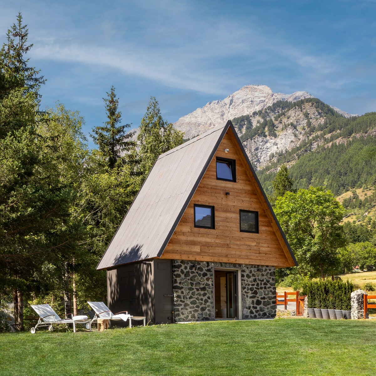 A modern chalet with a distinct a-frame design is set against a backdrop of mountains and greenery. The structure features a combination of stone and wooden elements. Sun loungers are arranged in the front yard, inviting relaxation in the surrounding natural landscape.