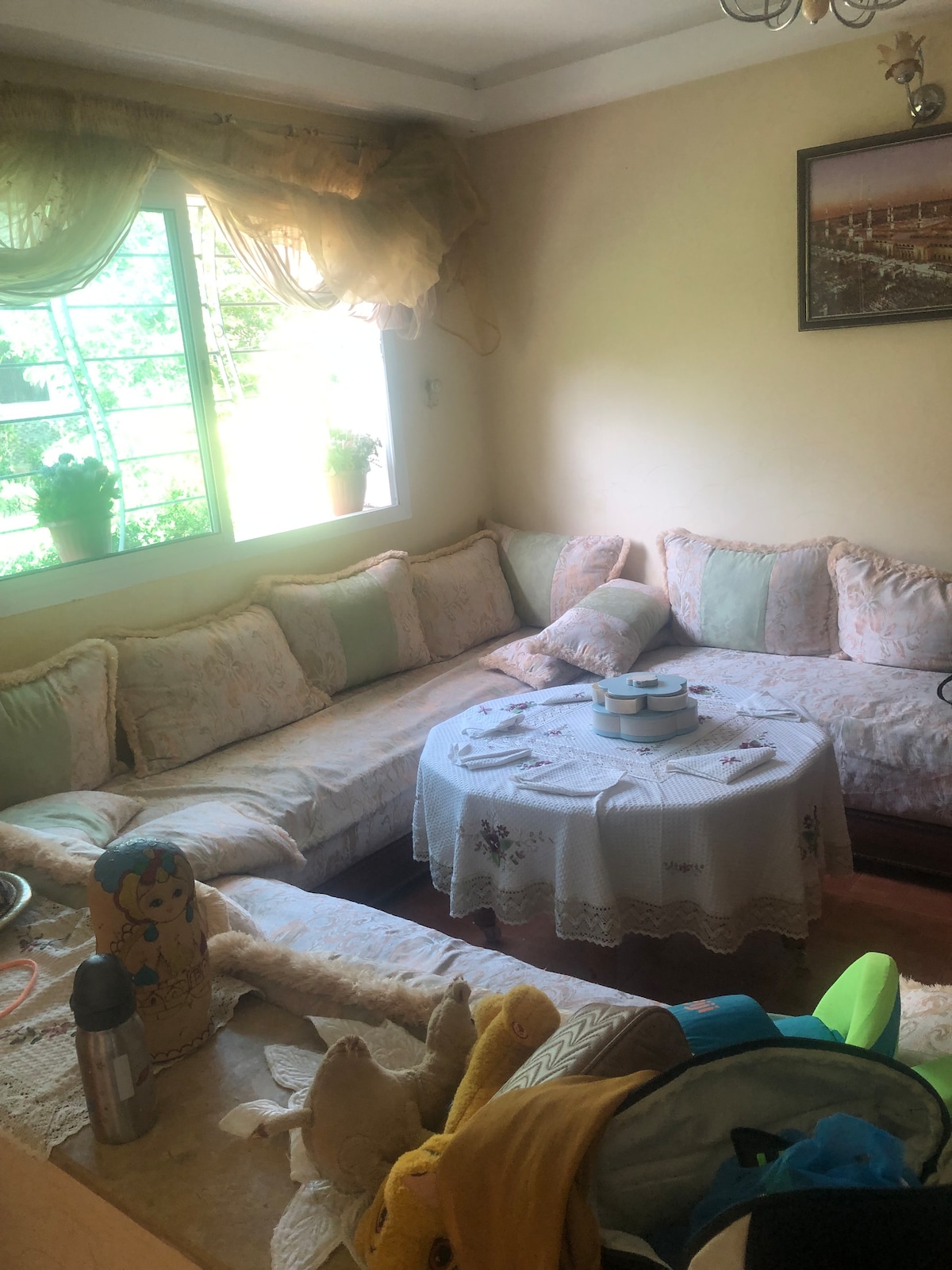 A comfortable seating area features a large, cushioned sofa adorned with soft pillows. A round table with a lace cover sits at the center, complemented by a small assortment of decorative items. Natural light enters through a window adorned with light drapes, enhancing the cozy ambiance.