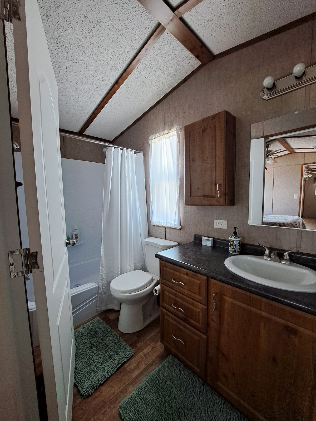 The bathroom layout features a shower and bathtub combination, a toilet, and a vanity with a sink. Natural light enters through a window covered by sheer curtains. Light green bath mats are placed on the floor, complementing the wooden cabinetry and neutral wall tones.
