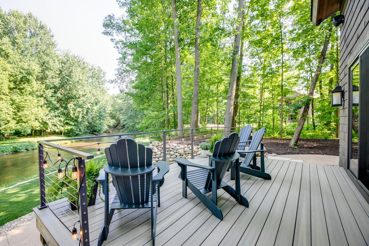 A tranquil deck overlooks the Dowagiac River, featuring several Adirondack chairs arranged for relaxation. Lush green trees provide a natural backdrop, creating a peaceful outdoor space. The area is designed for comfort, with a railing that enhances views of the surrounding landscape.