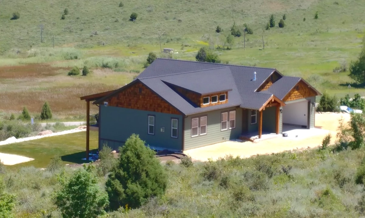 The exterior of the retreat is showcased, featuring a combination of stone and wood siding beneath a dark roof. The property is surrounded by vibrant greenery and rolling hills, with a spacious front yard leading to a covered entrance.