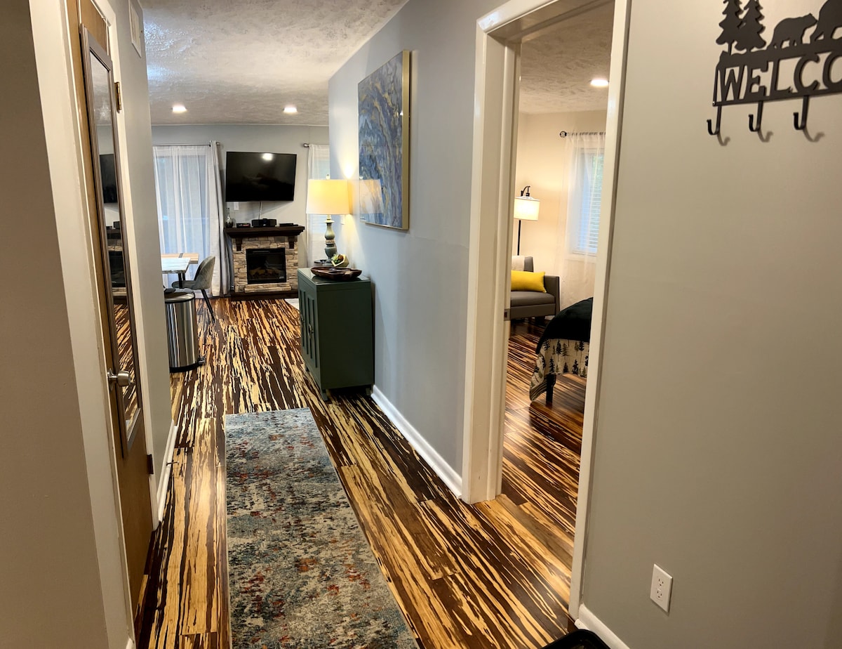 The hallway is lined with unique striped wooden flooring, leading to cozy living spaces. A small table with a lamp and decorative artwork adds warmth. In the distance, a living area features a fireplace and a TV, with natural light coming through the windows.