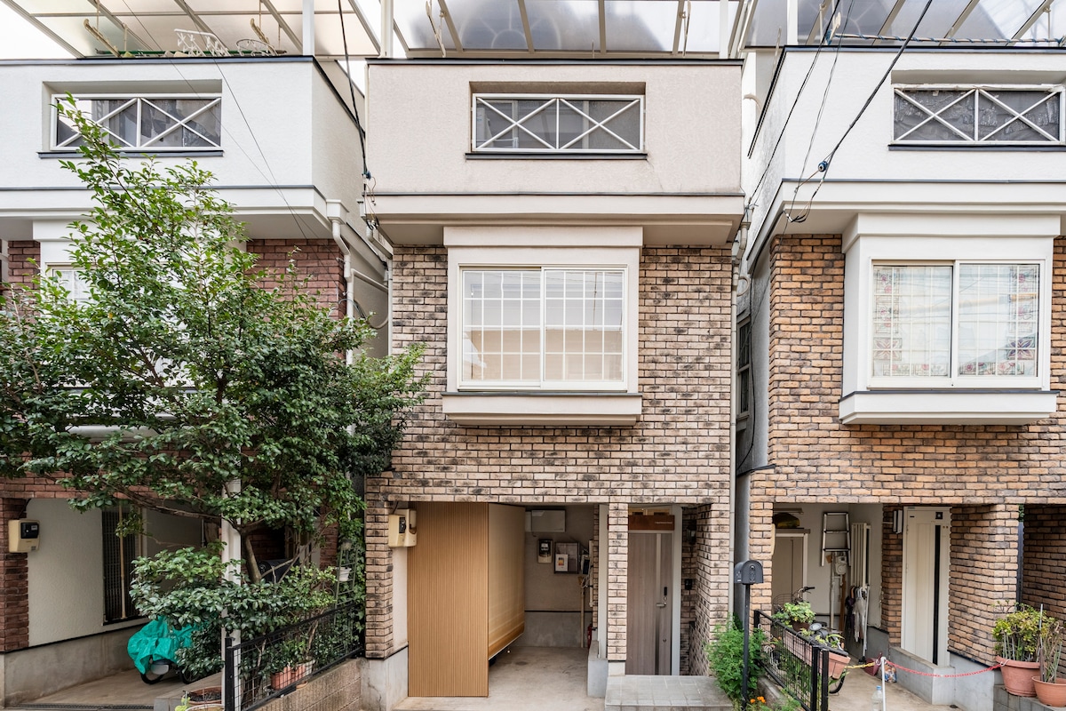 The exterior of a multi-story building is showcased, featuring a combination of brick and smooth facades. Large windows allow natural light into the interior, and a landscaped area with greenery is visible at the front. The structure adheres to a modern aesthetic.