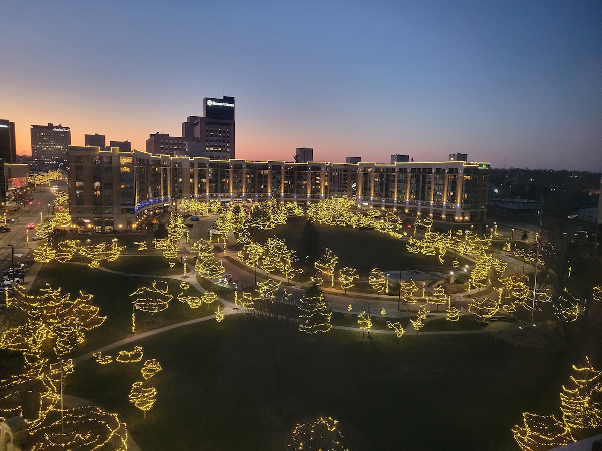An elevated view captures a park adorned with bright, twinkling lights along numerous trees, creating a festive atmosphere. The surrounding buildings are visible, while a gradient sunset sky showcases soft hues of blue and orange, adding depth to the serene scene.
