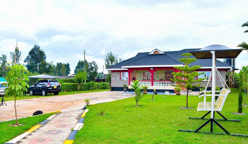 A spacious house is set within a verdant landscape, featuring a well-maintained lawn and a paved driveway. A shaded seating area is visible alongside the house, complemented by neatly trimmed plants and trees providing a refreshing ambiance.