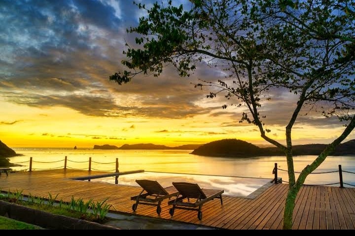 A serene sunset view over a calm water body, framed by a silhouette of a lush tree. Two lounge chairs are positioned on a wooden deck, inviting relaxation. An infinity pool reflects the colorful sky, enhancing the overall tranquility of the setting.