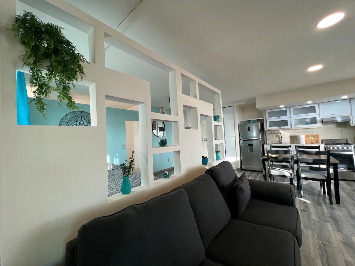 A modern living space features a dark gray sectional sofa, complemented by a decorative partition with built-in shelves. The shelves display turquoise accents. An adjacent kitchen area is visible, equipped with stainless steel appliances and contemporary cabinetry.