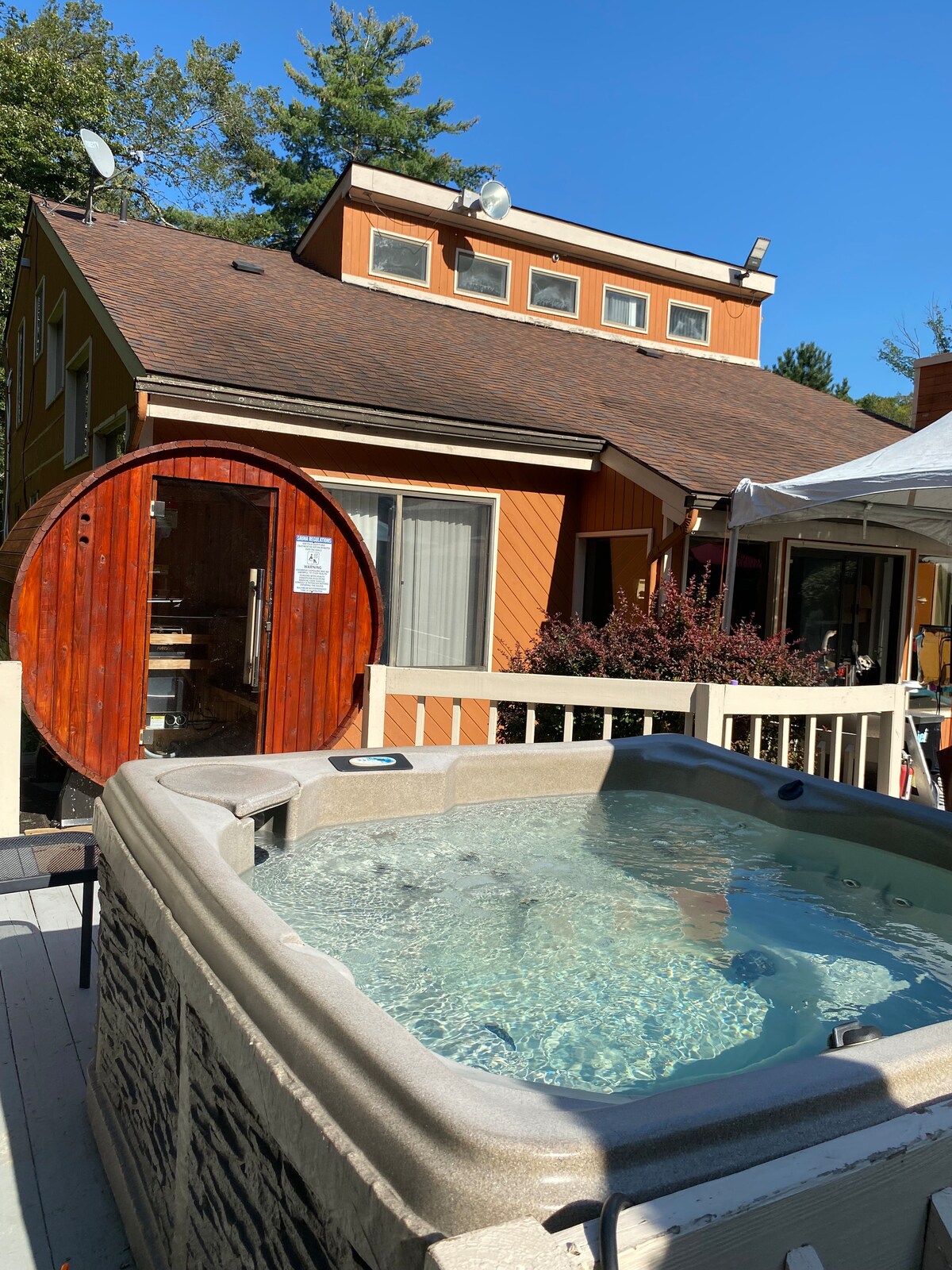 A hot tub is positioned on a wooden deck, featuring clear water and integrated seating. In the background, a colorful house is visible, alongside a circular wooden sauna. Surrounding greenery and a clear blue sky enhance the outdoor setting.