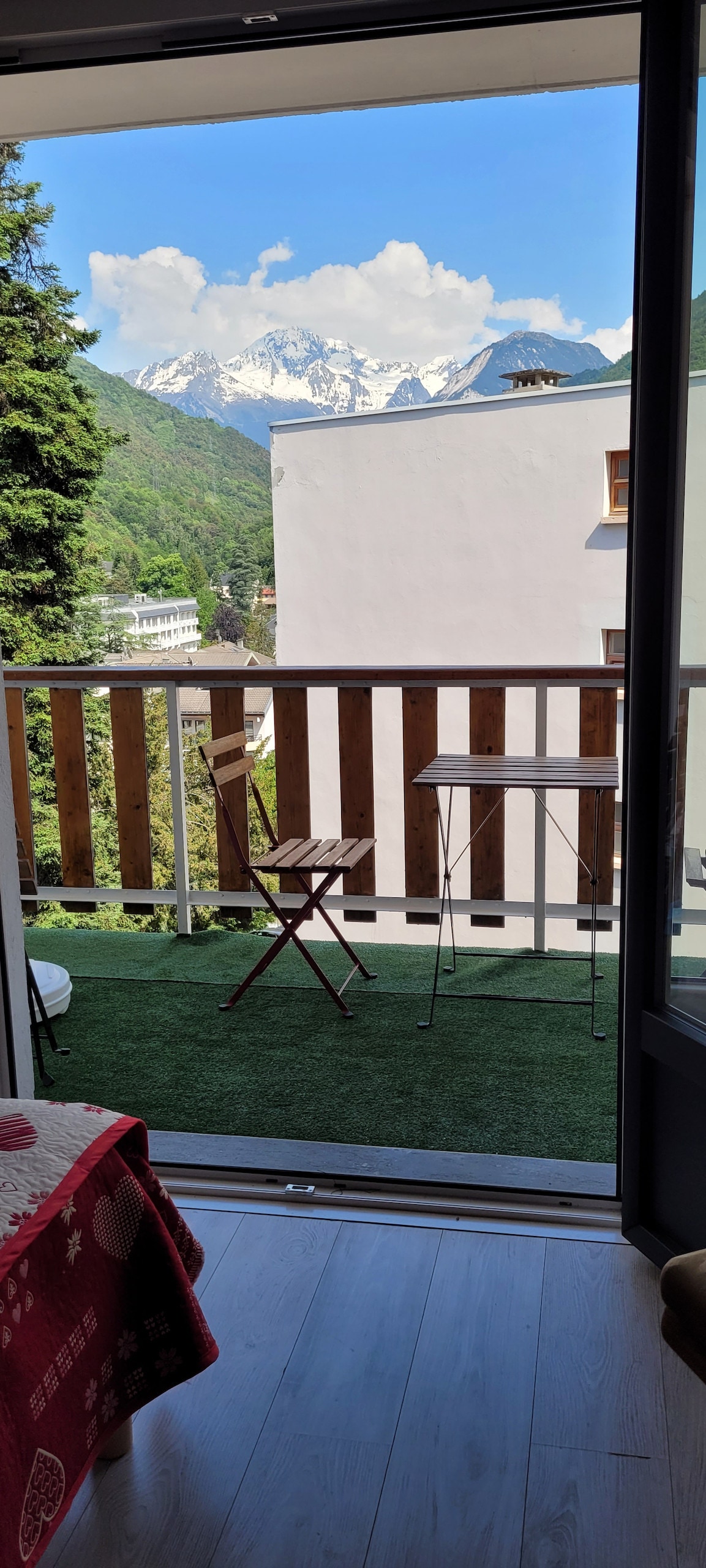 A view of a small outdoor balcony is presented, featuring a wooden table and chair set. The surrounding mountains are visible in the background, partially covered with clouds. Lush greenery is seen nearby, enhancing the serene setting.