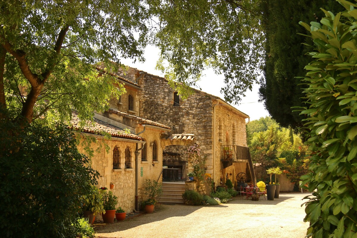 A traditional stone building is framed by lush greenery, showcasing a welcoming entrance with potted plants. Steps lead to the main door, while soft sunlight highlights the rustic architecture, creating a serene setting amidst the natural surroundings.