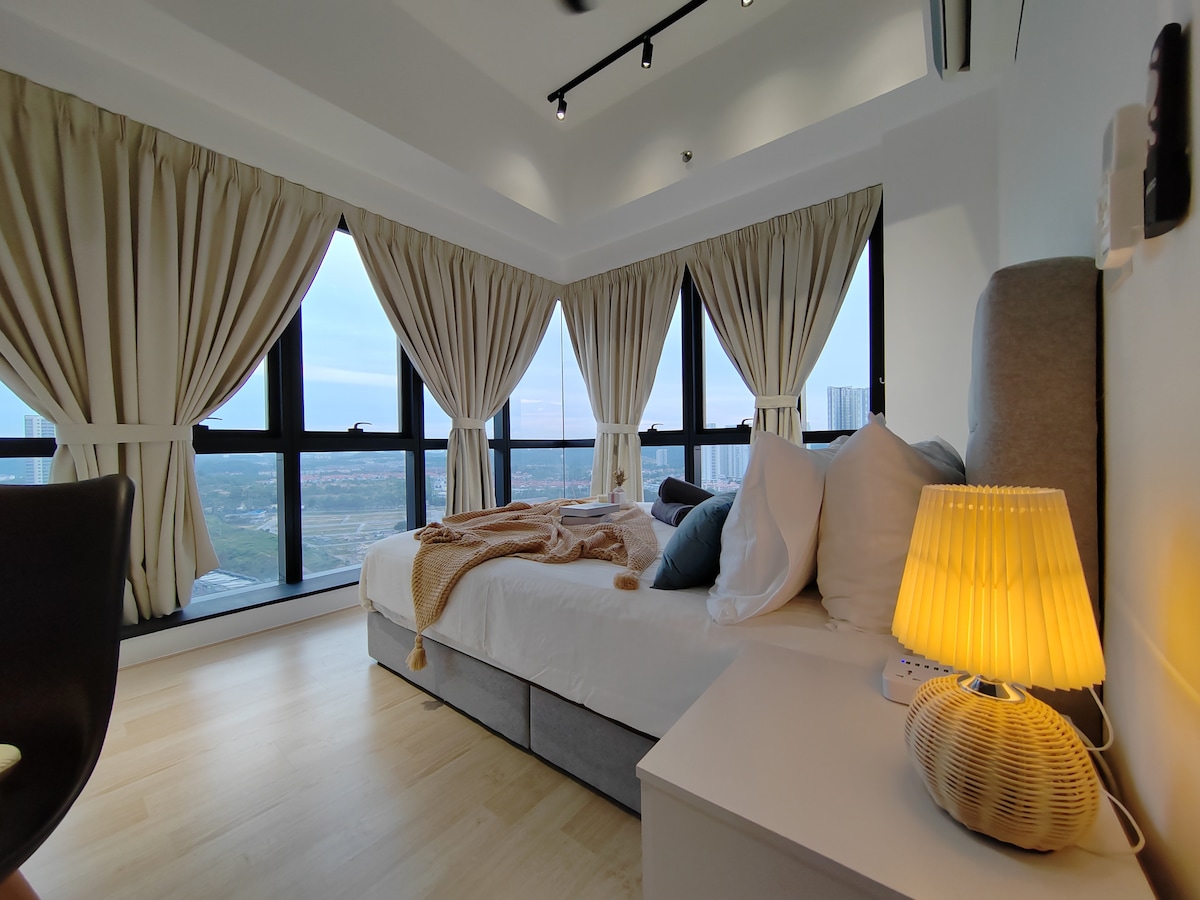 A well-appointed bedroom features a spacious bed with soft pillows and a warm throw. Large windows are framed by elegant curtains, providing natural light and a scenic view. A bedside lamp adds a touch of warmth, accompanied by a modern nightstand.