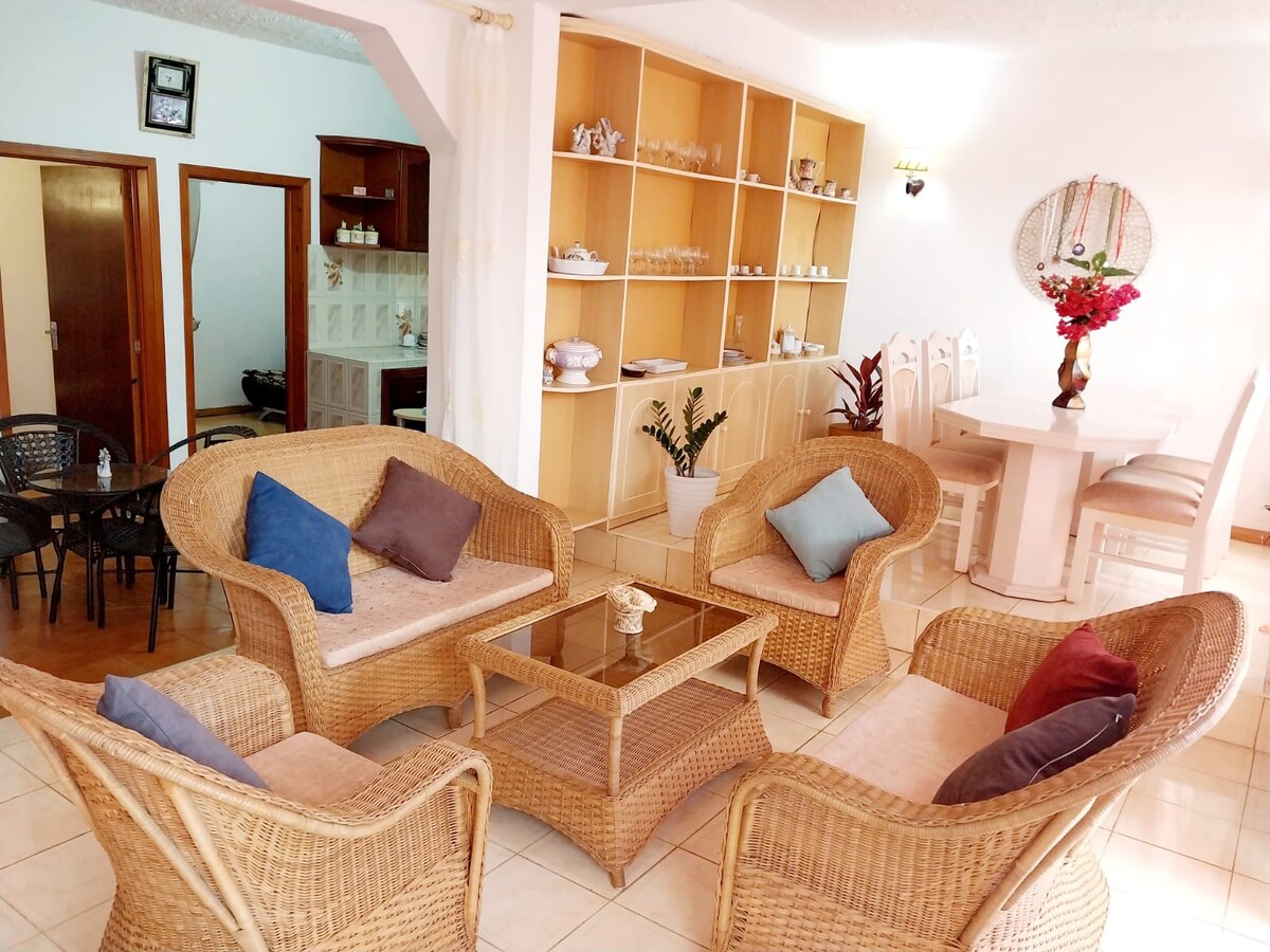 A living area features woven seating arranged around a glass-topped coffee table. Decorative cushions in various colors are placed on the chairs. Light-colored tiles extend across the floor, and built-in shelving displays dishes and decor items in the background.