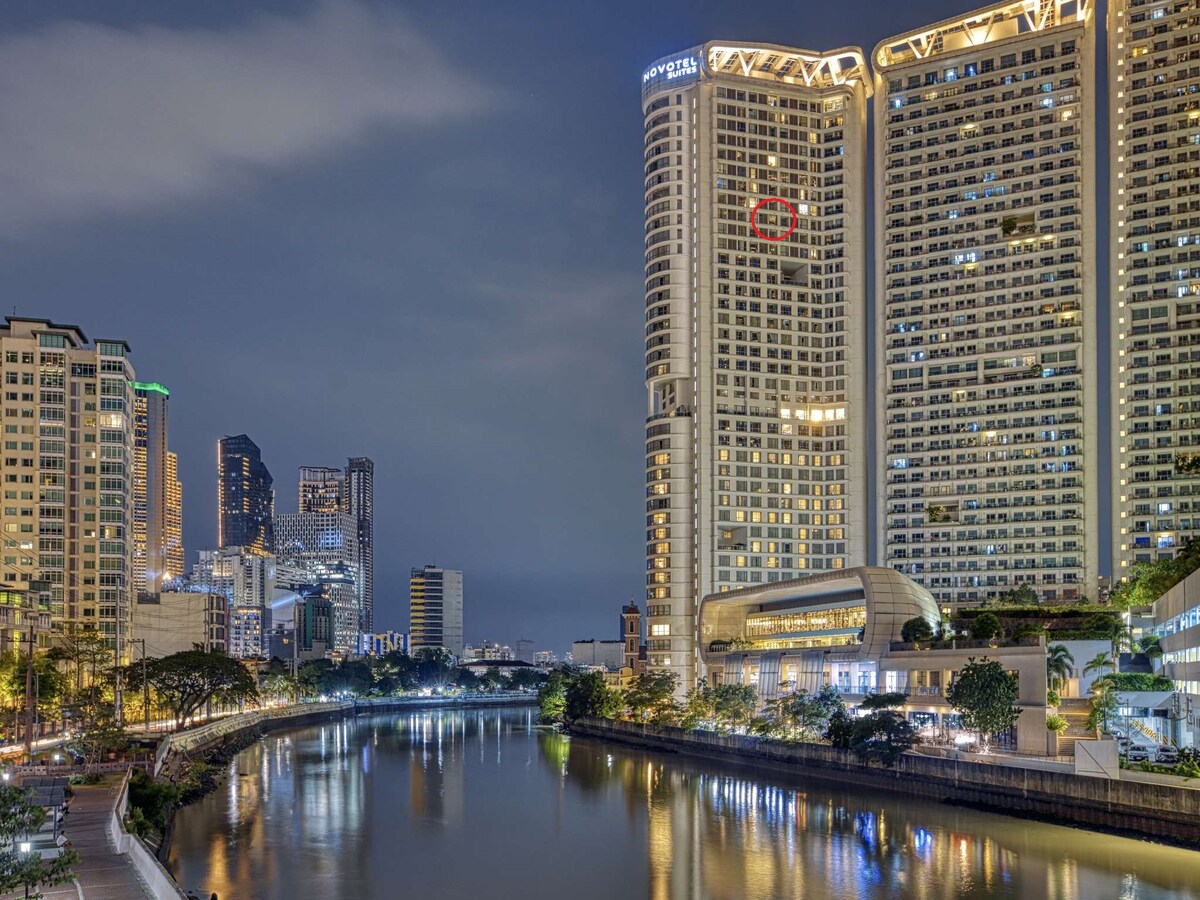 The image showcases a panoramic view of the Pasig River at night, framed by the illuminated skyline of Manila. The Novotel Tower at Acqua Private Residences stands prominently, with its distinct architecture reflecting the vibrant city lights against the serene waters.