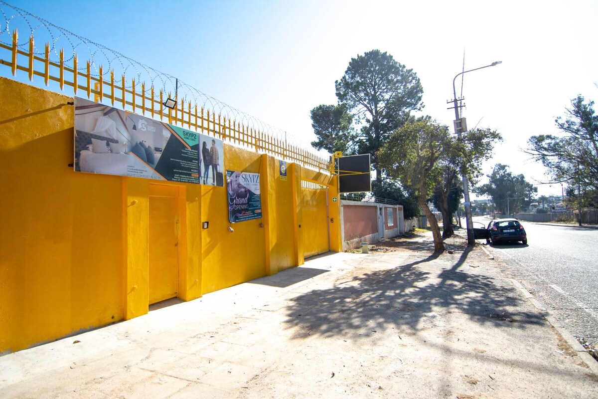 The exterior of a bright yellow building is showcased, featuring a secure entry with barbed wire atop the fence. Nearby, trees provide some shade, while a parked car is seen along the quiet street. The clear sky adds to the vibrant appearance of the setting.