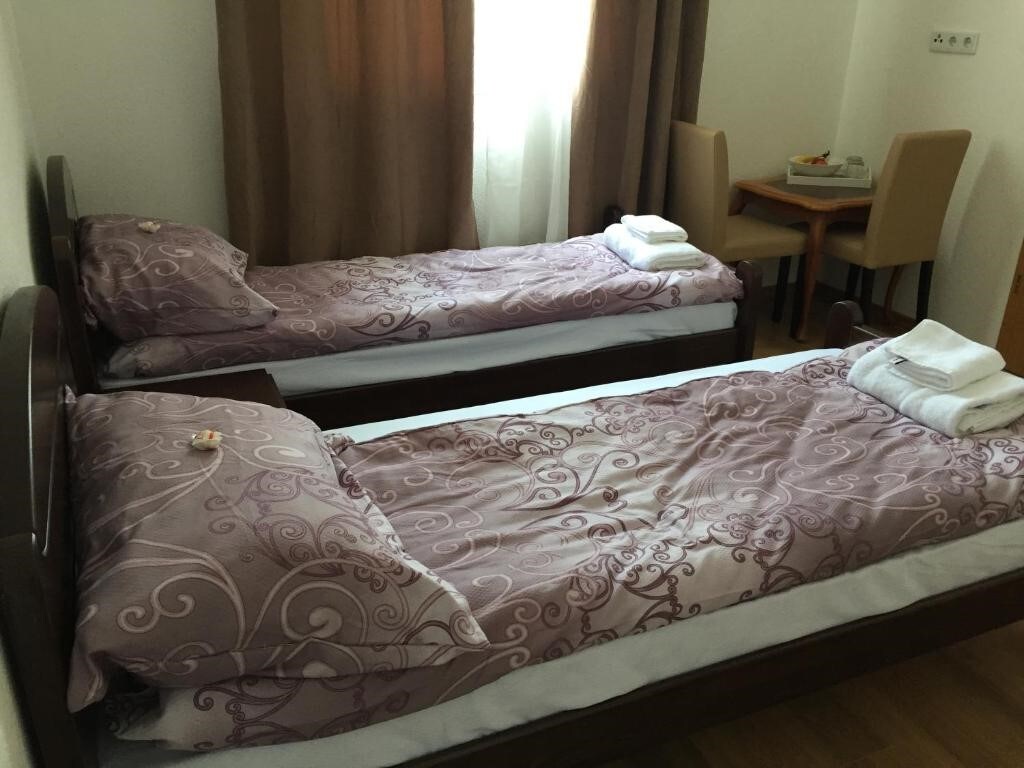 The image displays a cozy sleeping area featuring two separate beds, each adorned with patterned comforters and matching pillows. Soft, muted curtains allow natural light to fill the space. A small dining table with two chairs is visible in the background.
