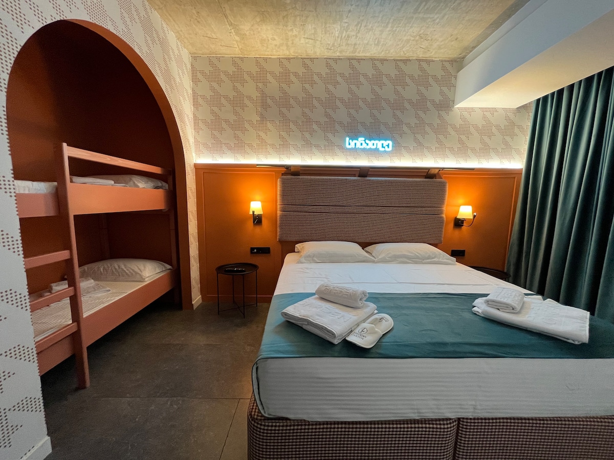 A sleeping area features a spacious bed with fresh linens, complemented by a decorative headboard. A cozy bunk bed is positioned against the wall. Soft lighting enhances the room's design, while a side table adds functionality.
