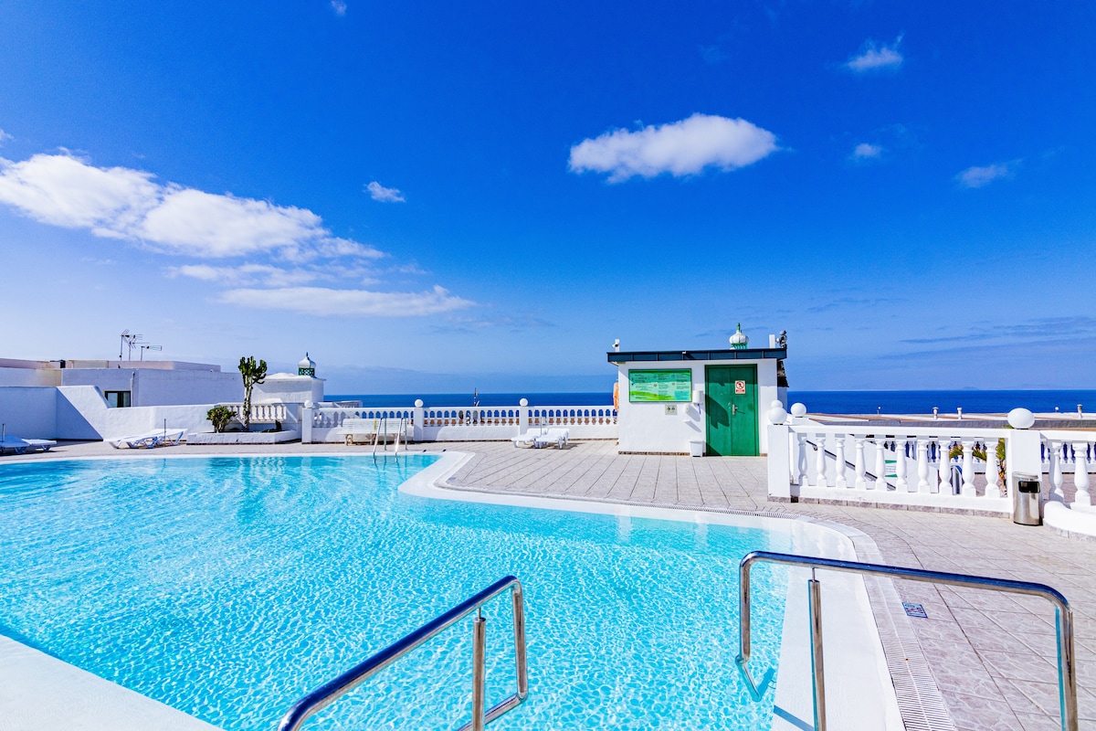 A clear blue sky reflects over a communal swimming pool, with bright turquoise water inviting relaxation. Surrounding areas are paved with grey tiles and include white railings and a small green building in the background, enhancing the open and airy feel of the space.