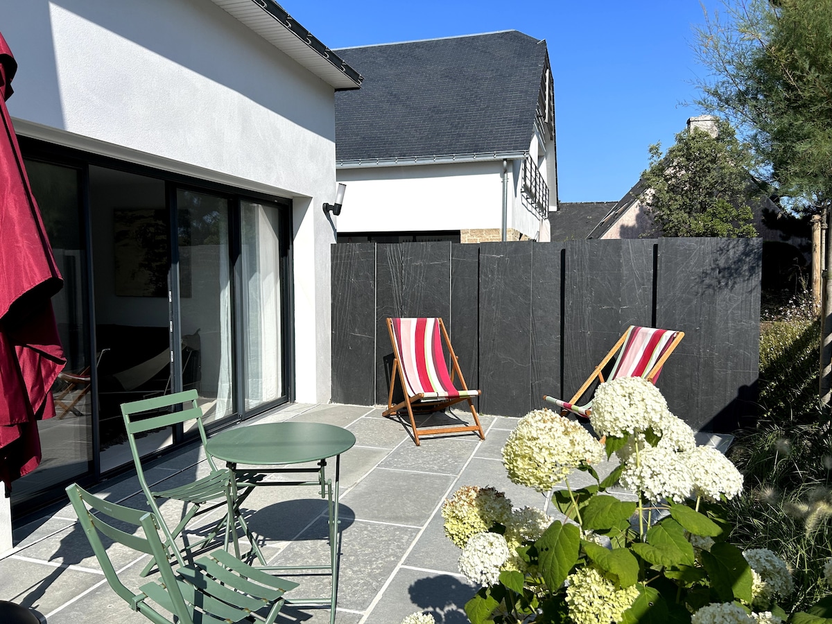 An outdoor patio area is featured, with a round table and four green chairs positioned on gray tiles. Two lounge chairs with striped fabric are visible, along with flowering plants that add greenery to the space. A wall provides privacy, and clear blue skies are above.