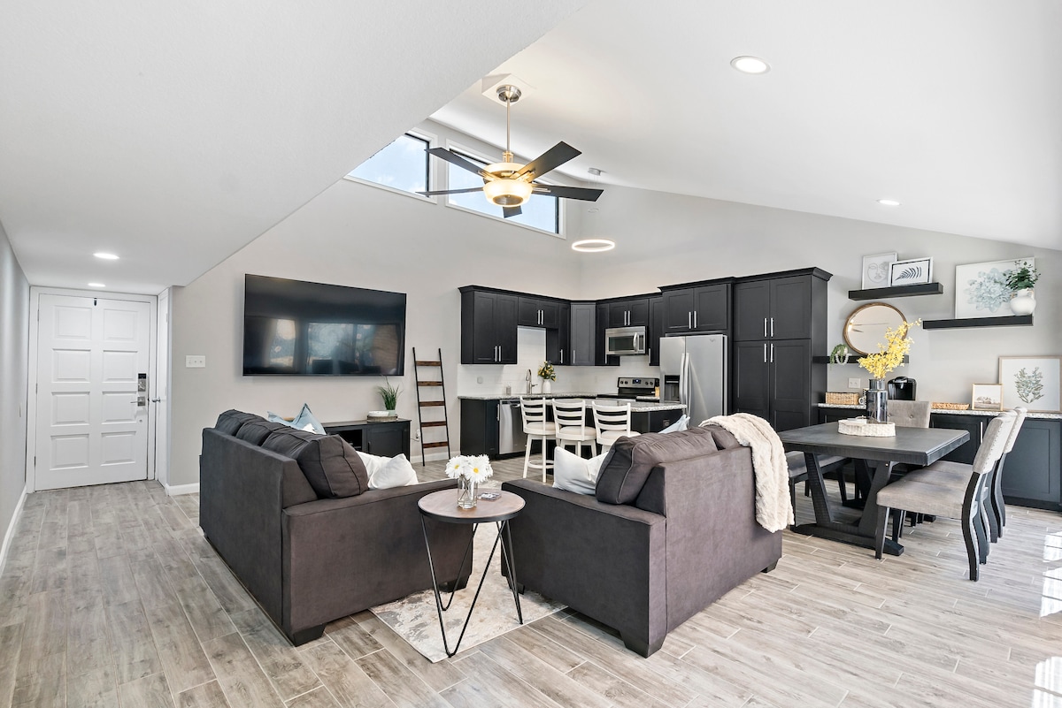 An open living area features a spacious layout with a modern kitchen, dining table, and comfortable seating. Natural light floods through a skylight, illuminating the dark cabinetry and light-colored flooring. A decorative ladder leans against the wall, adding a touch of elegance.