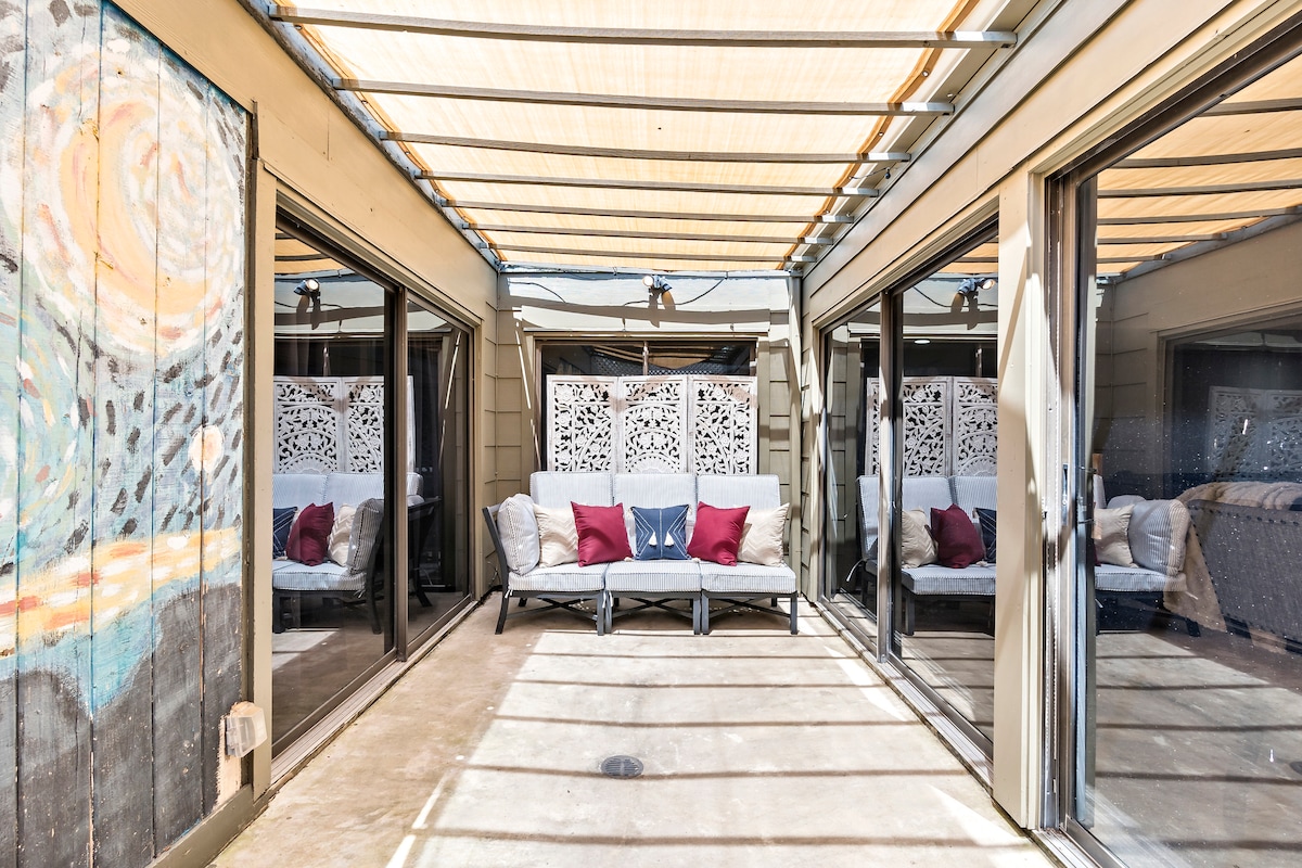 A screened-in patio area is depicted, featuring two cushioned couches with decorative pillows. Large sliding glass doors provide natural light and an open feel, while a patterned privacy screen adds texture to the backdrop. The ceiling is adorned with fabric awnings, offering shade.