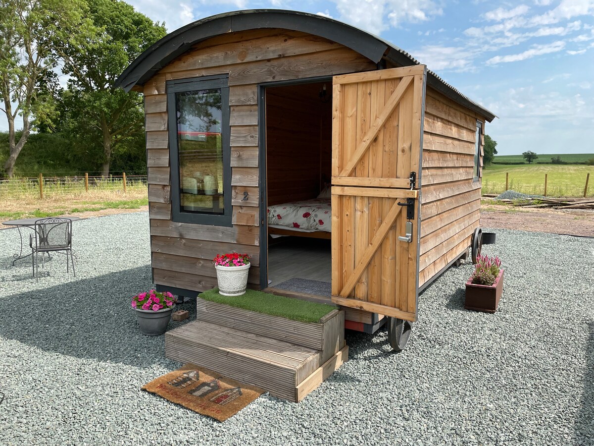 A wooden shepherd's hut with an open door reveals a cozy interior. The exterior features a rustic finish, surrounded by gravel and planters with flowers. A small outdoor seating area with two chairs is visible nearby, set against a backdrop of green fields.