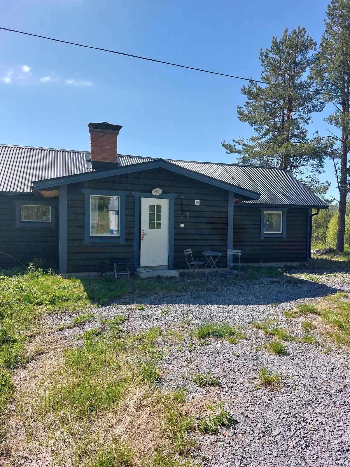 The charming exterior of the cabin is shown, featuring a blue wooden façade and a welcoming front door. A small outdoor seating area with two chairs and a table is positioned beside the entrance, surrounded by lush greenery and gravel.