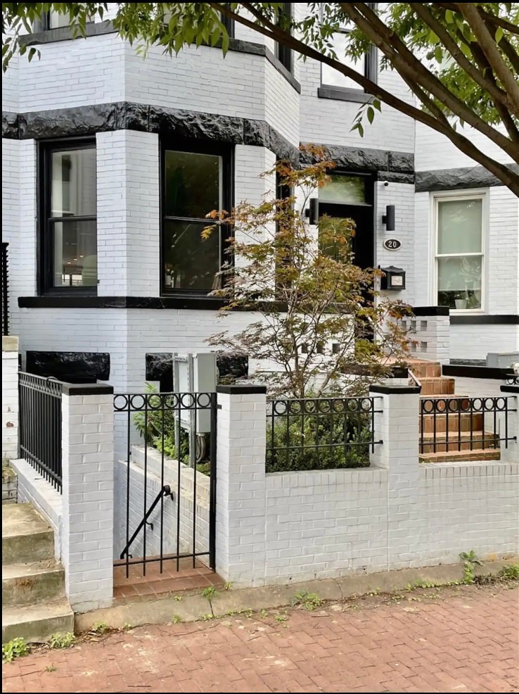 A classic Victorian row-home is shown with a white brick exterior and black trim. A small decorative tree is visible next to the entrance steps, which lead to a black door framed by large windows. A white fence surrounds the property, providing a quaint, welcoming appearance.