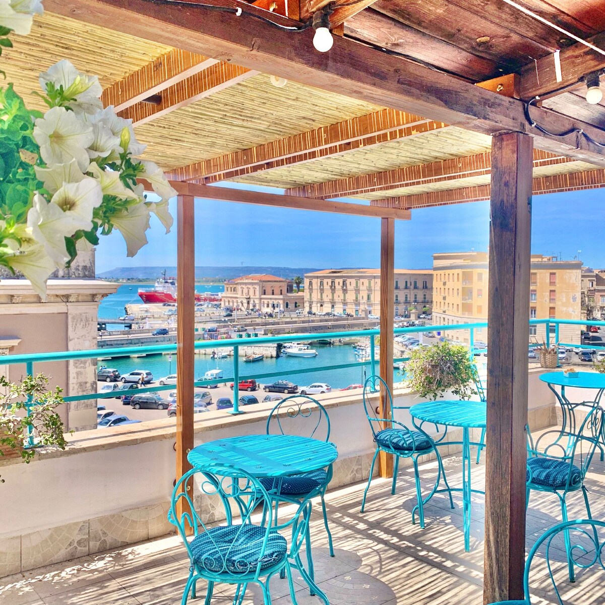A terrace features turquoise metal tables and chairs, surrounded by potted greenery. The view reveals a bustling marina with boats docked in the water, complemented by historic buildings in the background under a clear blue sky.