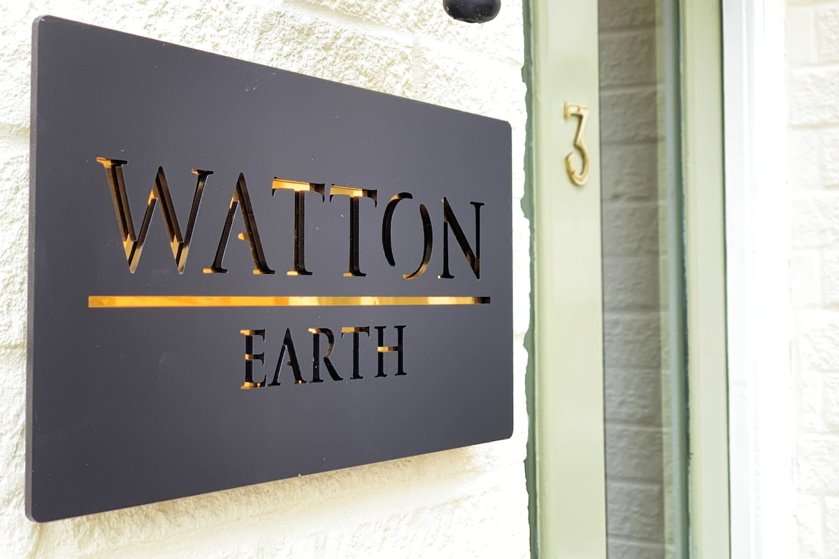 A modern sign reading 'WATTON EARTH' is affixed to a textured white wall, showcasing sleek black lettering with a gold accent. The entrance is complemented by a green door with the number '3' displayed in a minimalist style beside the sign.