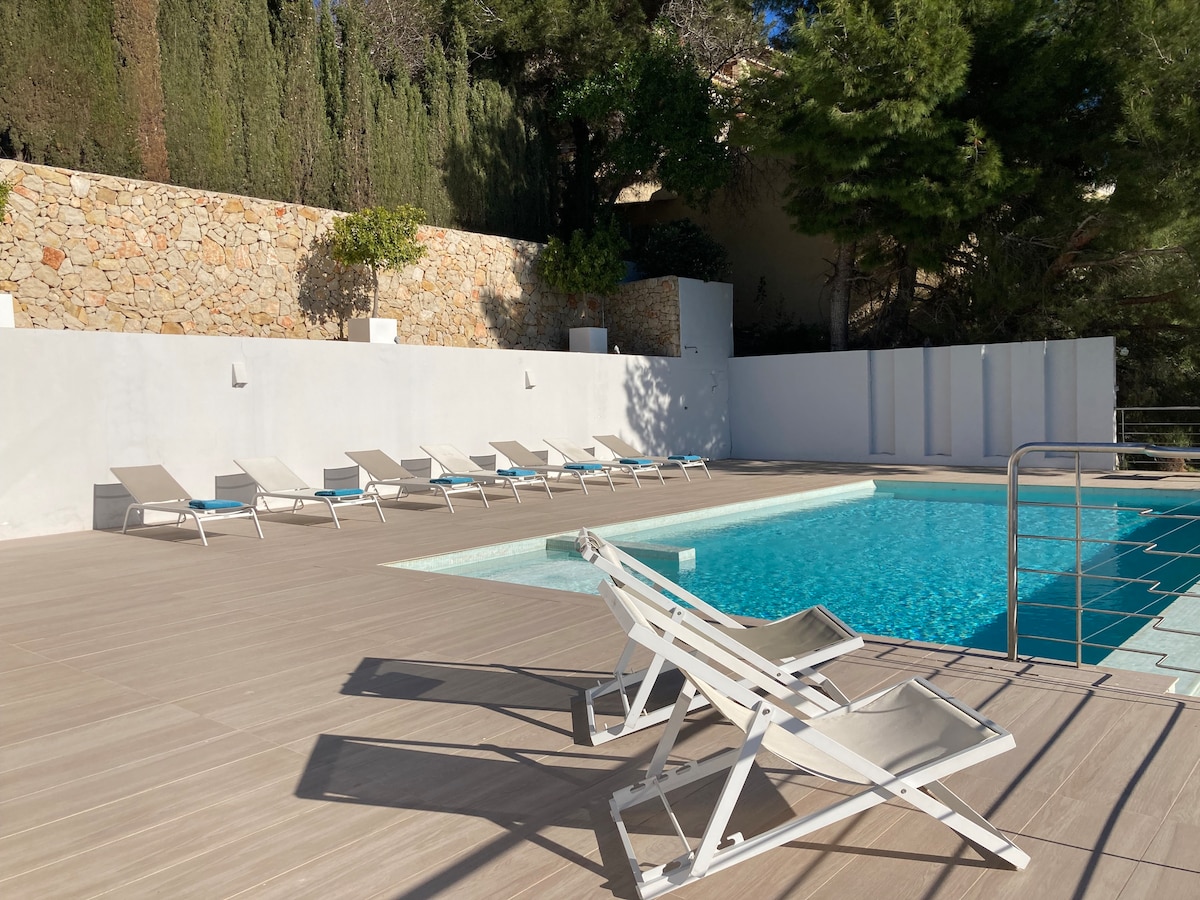 A calm outdoor area features a private swimming pool surrounded by a spacious wooden deck. Multiple sun loungers are arranged neatly along the pool, with stone walls and lush greenery providing a peaceful backdrop.