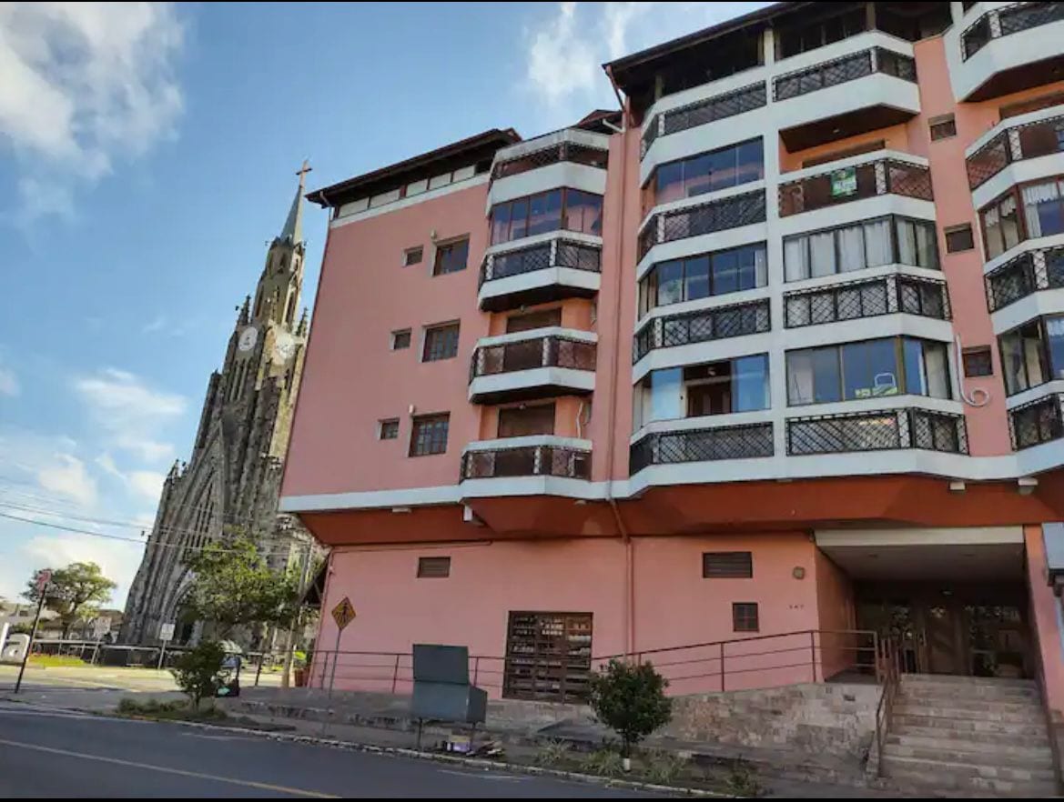 The building features a distinct pink facade with multiple balconies, showcasing a variety of window styles. The iconic stone church stands prominently beside the structure, adding character to the scene. Clear skies and sunlight highlight the layout of the apartment's exterior.