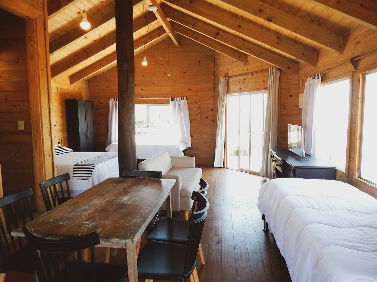 A spacious wooden interior features two beds and a comfortable sofa, creating a relaxed atmosphere. Large windows allow natural light to fill the space, while a dining table with black chairs is positioned adjacent. Dark curtains frame the windows, enhancing privacy.