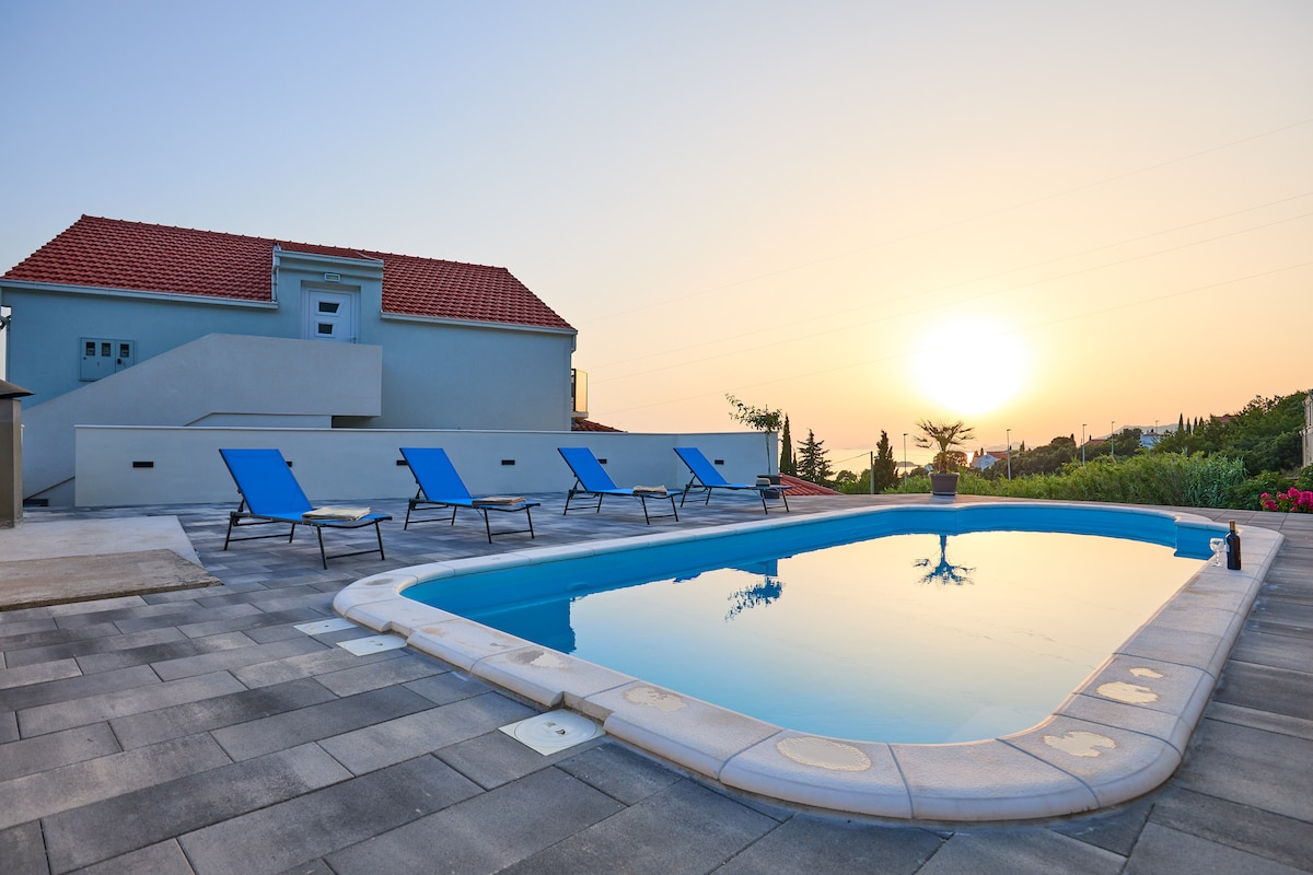 A private pool area features several blue lounge chairs arranged around a clear water pool. The tranquil setting is complemented by a serene sunset, casting warm hues over the surrounding landscape and the modern structure in the background.