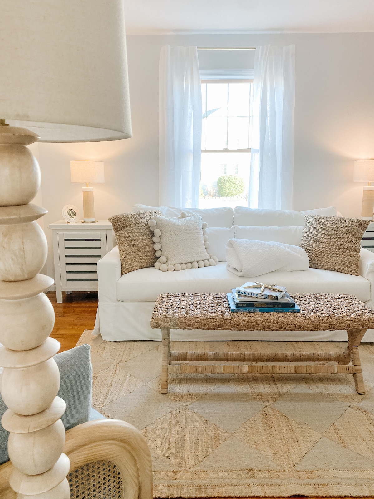 A light-filled living area features a white sofa adorned with textured pillows and a cozy throw. A woven coffee table sits in front, complemented by a neutral area rug. Soft lighting is provided by table lamps on either side, enhancing the room's calm atmosphere.