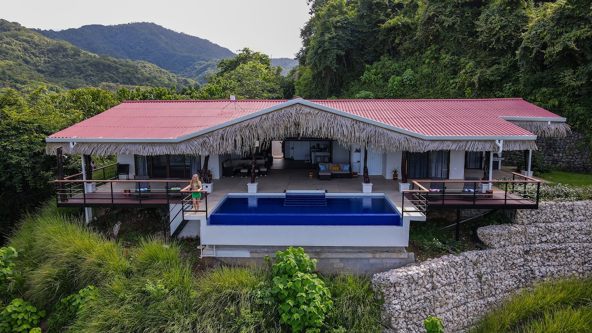 An aerial view of a modern villa showcases a striking infinity pool that blends into the horizon. The villa features a wide terrace with palm thatched roofs, surrounded by vibrant green hills and lush vegetation, providing a serene backdrop.