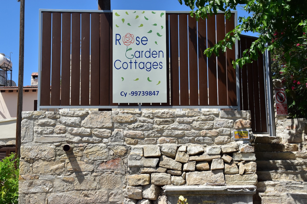 The entrance to Rose Garden Cottages is marked by a stone wall and a wooden fence. A prominently displayed sign features the cottage's name, along with contact information. Lush greenery surrounds the area, adding a natural touch to the welcoming façade.