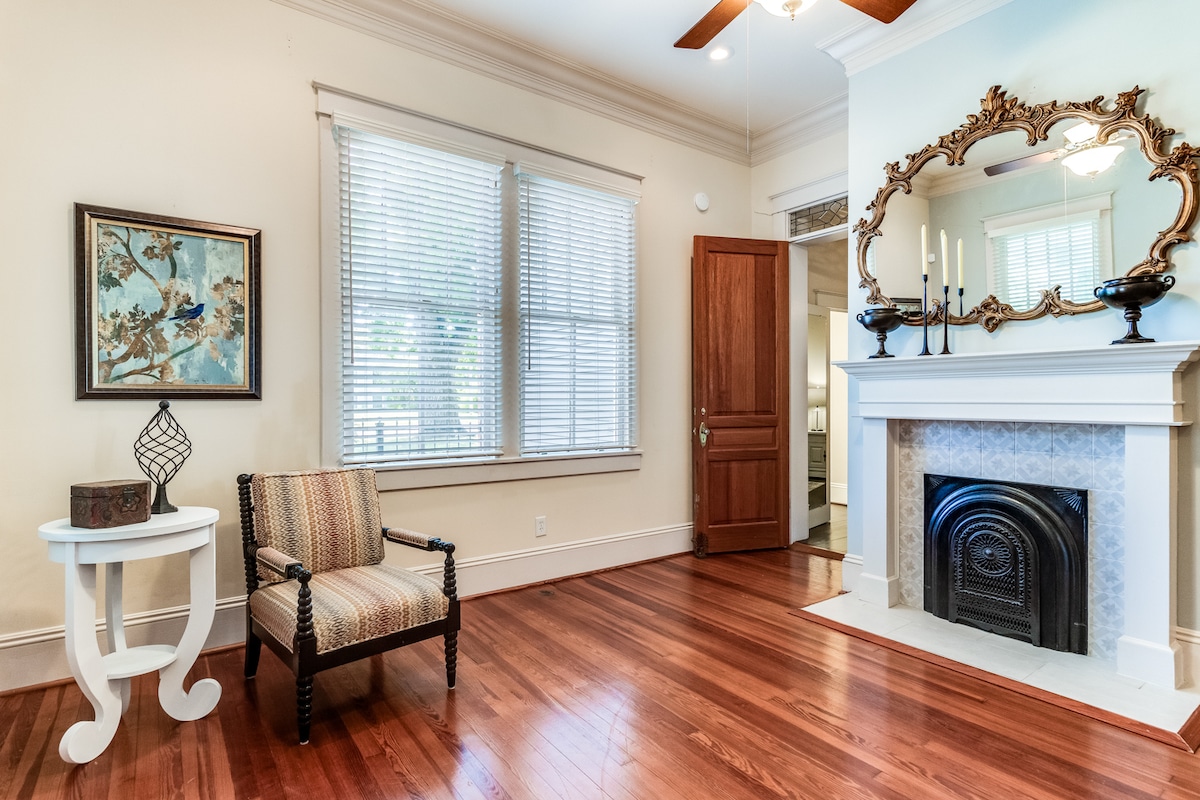 A welcoming living area is showcased with polished hardwood floors and natural light flowing through the window. A decorative mirror is mounted above a fireplace, while a small side table and a striped chair complete the inviting space.