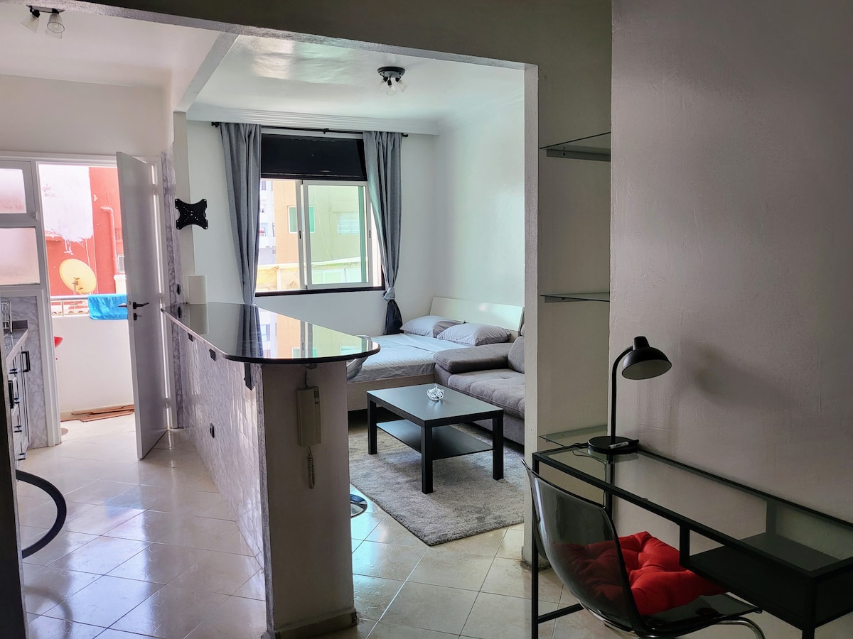 A studio apartment is displayed with a modern layout featuring natural light from a large window. A spacious bed is positioned beside a small coffee table, while a sleek desk with a lamp complements the decor. A bar area separates the kitchen from the living space.